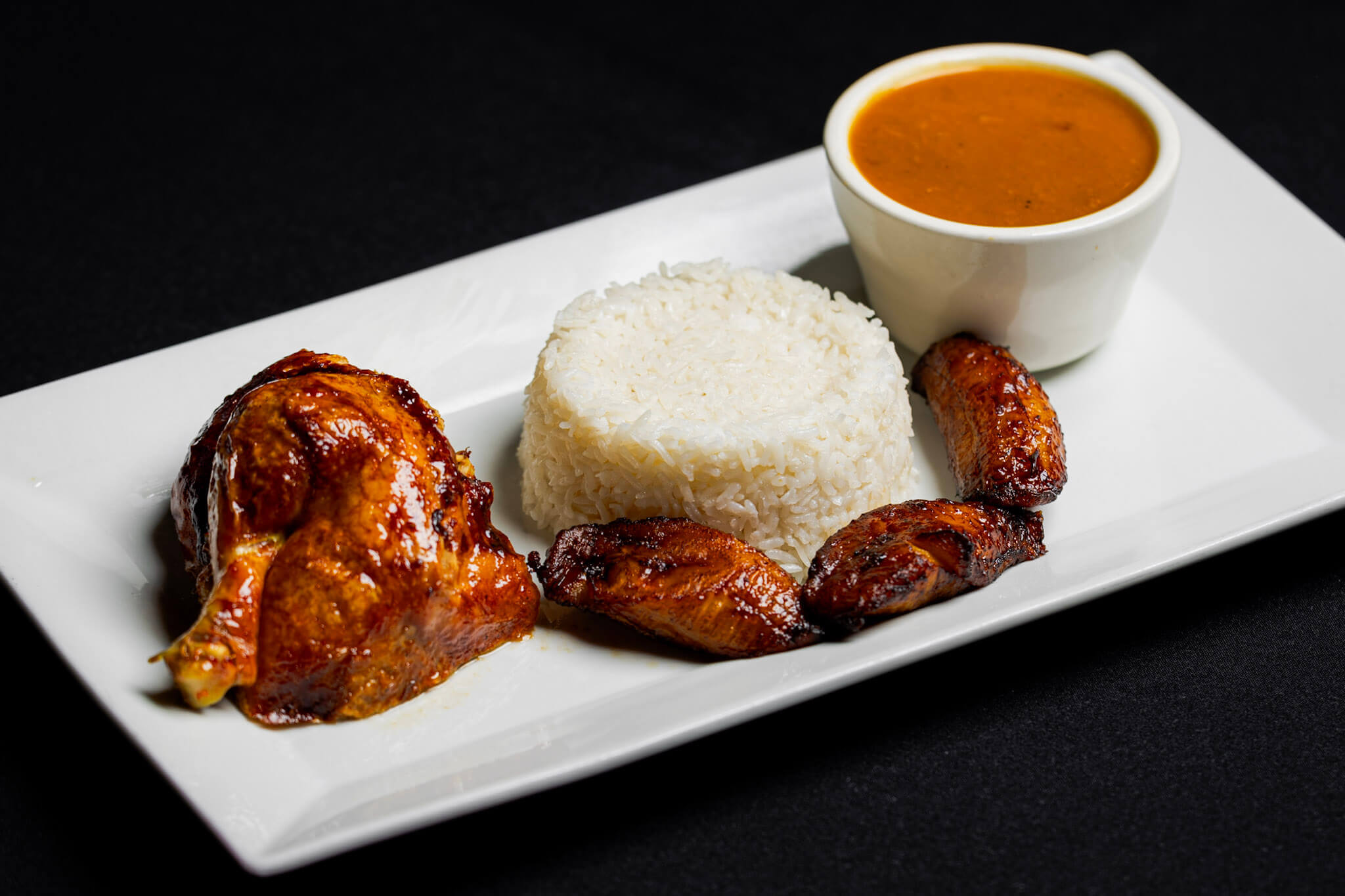 A plate with roasted chicken, a mound of white rice, fried plantains, and a cup of brown sauce on a white rectangular dish against a black background.