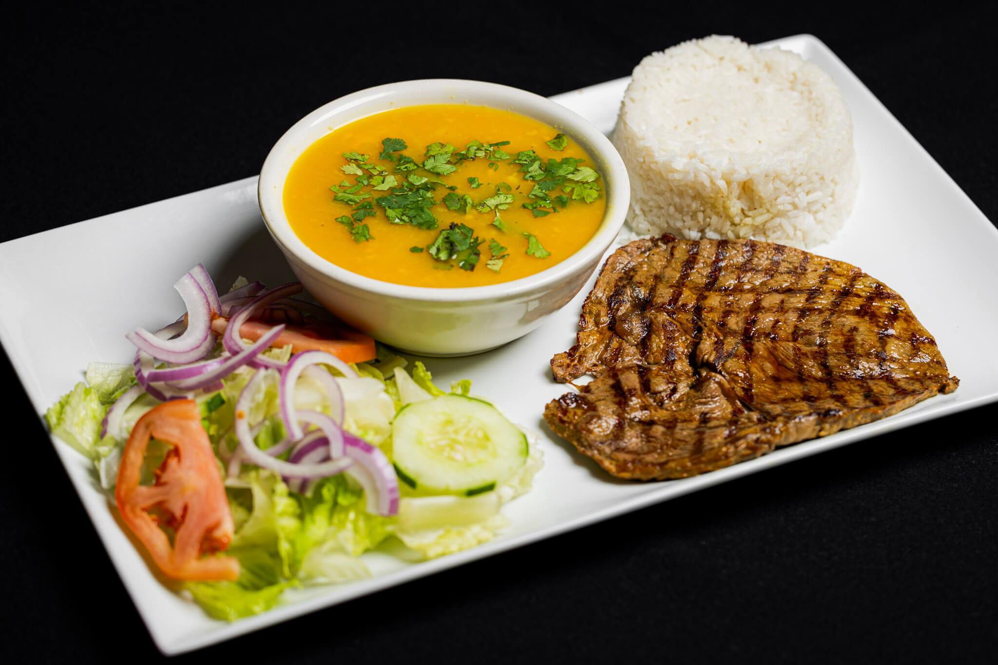 A plate with white rice, grilled steak, a bowl of soup garnished with herbs, and a salad of lettuce, tomato, cucumber, and red onion on a white rectangular dish.