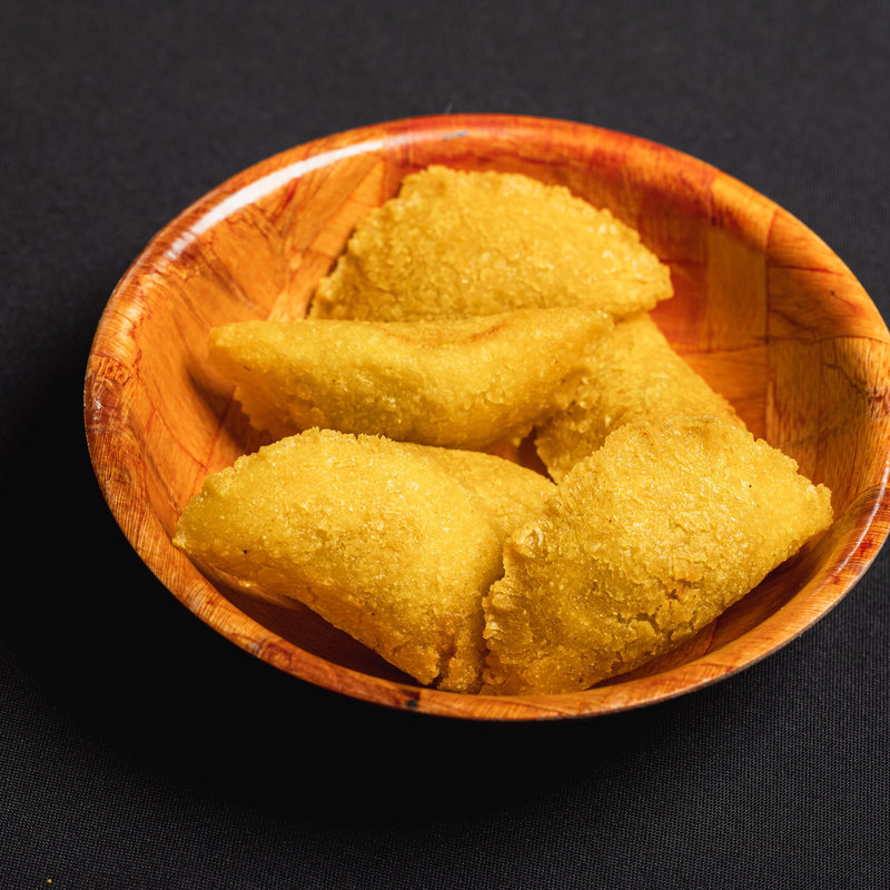A wooden bowl contains four fried, golden-brown, triangular pastries on a black surface.