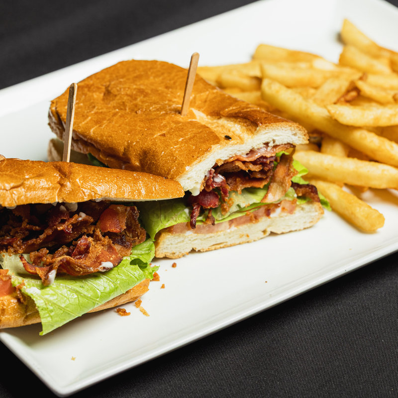 A sandwich with bacon, lettuce, tomato, and mayonnaise is cut in half on a white plate, served with a side of French fries.