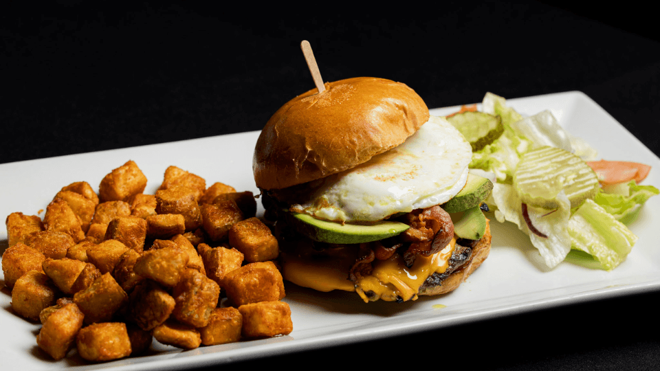 A cheeseburger with fried egg, bacon, and avocado, served with seasoned tater tots and a side salad on a rectangular white plate.