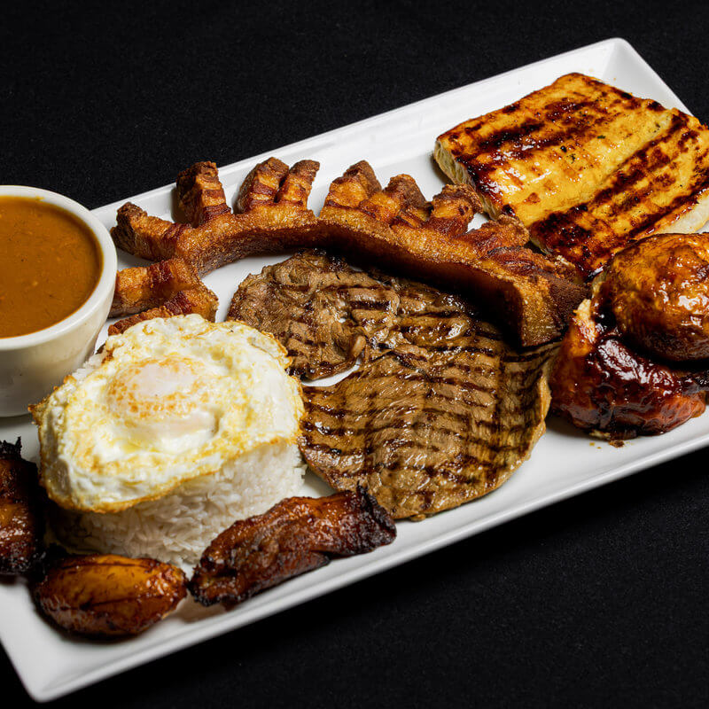 Rectangular plate with grilled beef, fried plantains, rice topped with a fried egg, pork crackling, grilled chicken, a slice of grilled cheese, roasted sausage, and a cup of brown sauce.