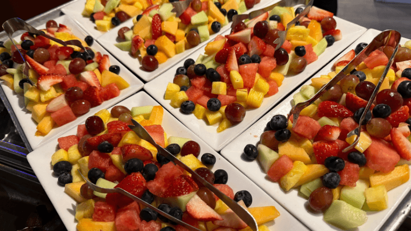 Six rectangular plates filled with assorted fresh fruit, including strawberries, blueberries, grapes, melon, and pineapple, with metal tongs on each plate.
