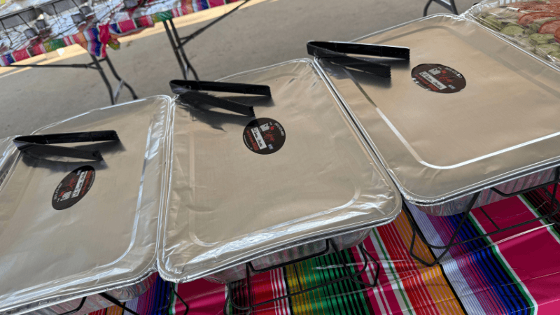 Three aluminum food trays with lids and black tongs rest on a colorful tablecloth at an outdoor event.