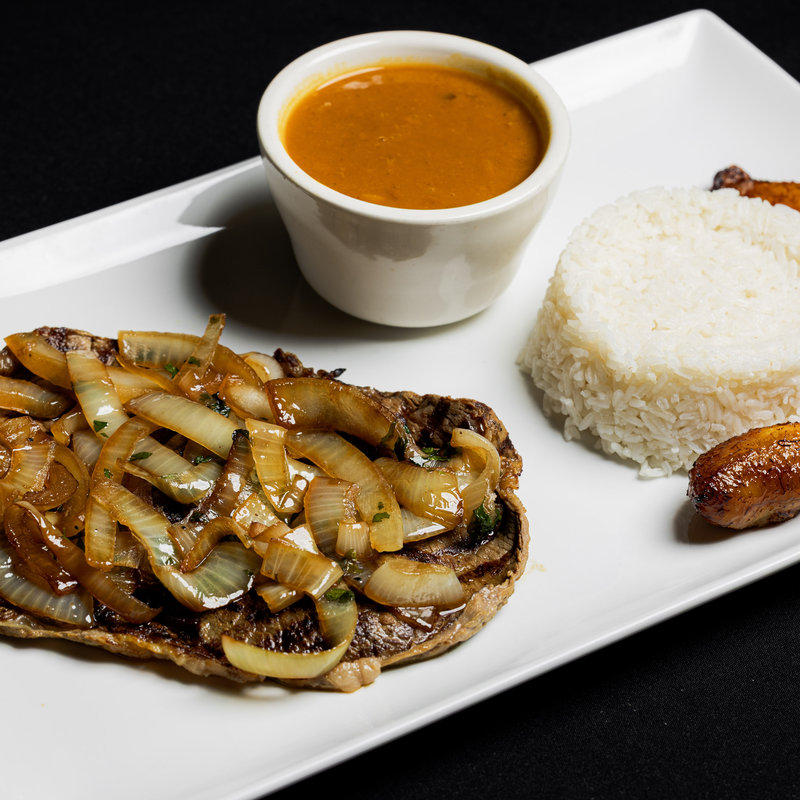 A plate with grilled steak topped with sautéed onions, white rice, fried plantains, and a cup of brown sauce on a rectangular white dish.