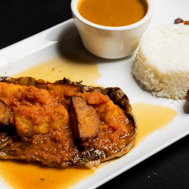 A plate with fish topped with tomato sauce and fried plantains, served with white rice and a small cup of soup on the side.