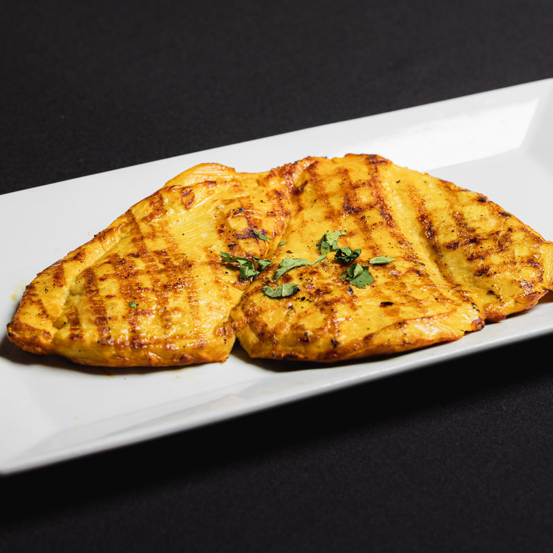Two pieces of grilled, golden-yellow chicken breast garnished with chopped herbs on a rectangular white plate, set against a black background.