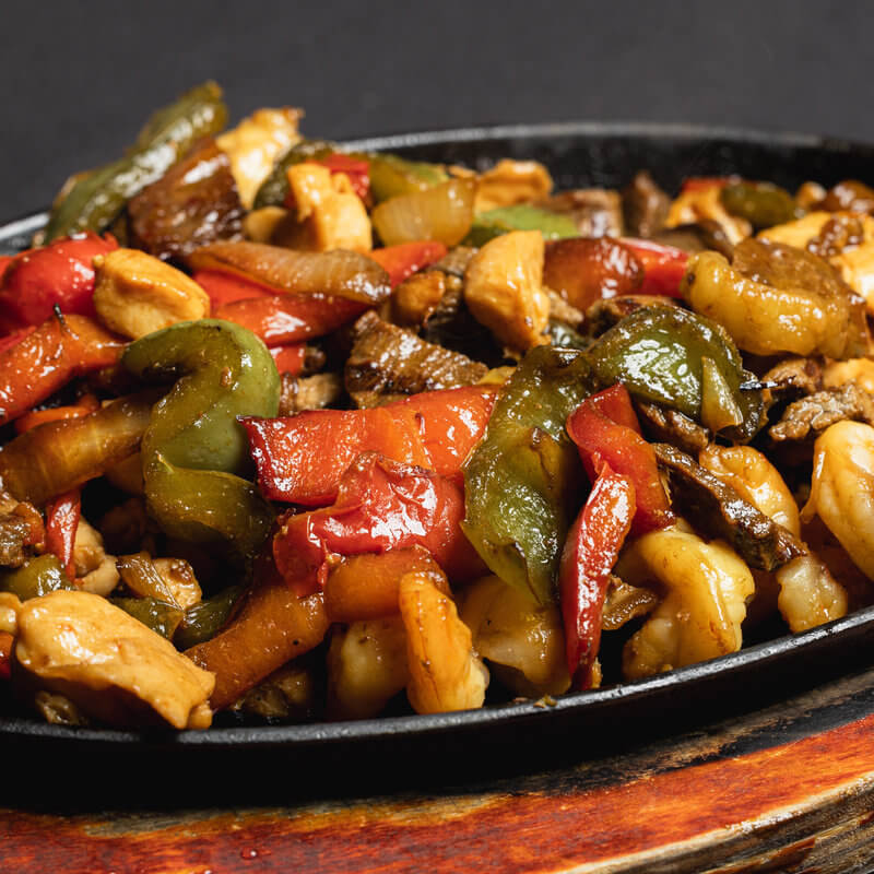 A sizzling platter of stir-fried chicken, beef, and vegetables including red and green bell peppers, served on a black dish.