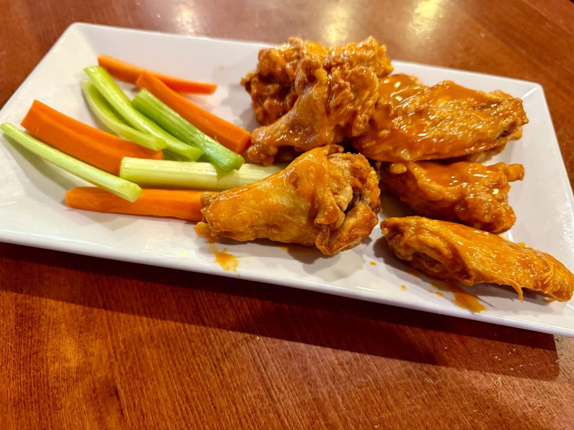Plate of buffalo chicken wings with celery and carrot sticks on a white rectangular dish, set on a wooden table.