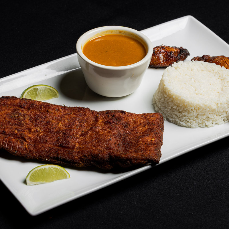 A rectangular plate with grilled meat, a mound of white rice, a cup of brown sauce, two lime wedges, and a piece of fried plantain, all on a black background.
