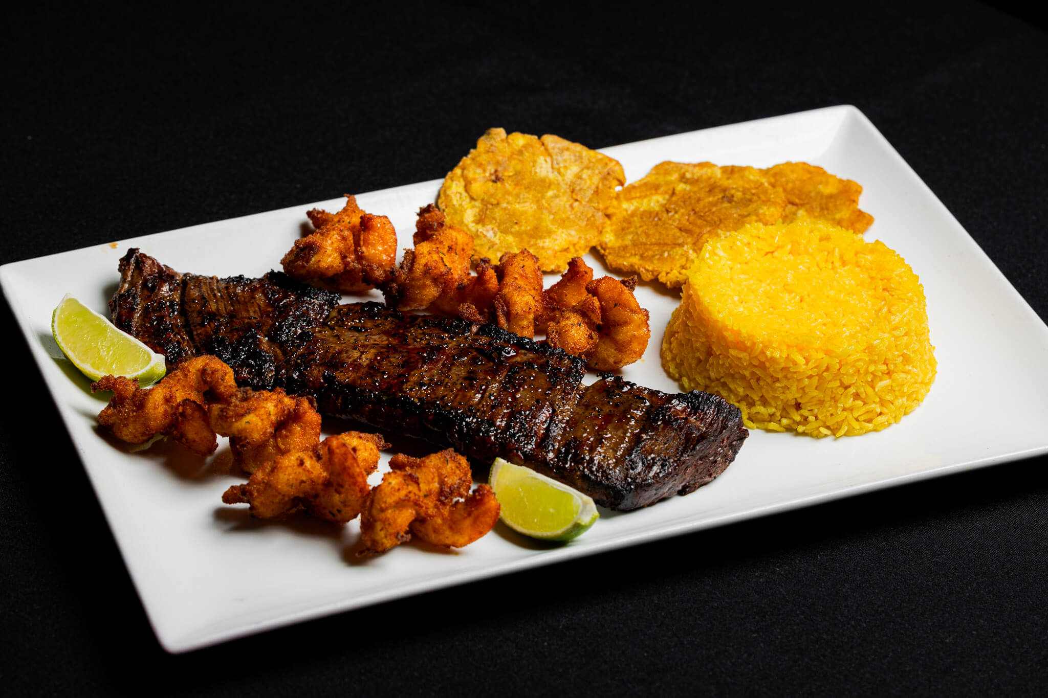 A rectangular white plate with grilled steak, fried shrimp, yellow rice molded into a round shape, two tostones, and lime wedges, set against a black background.