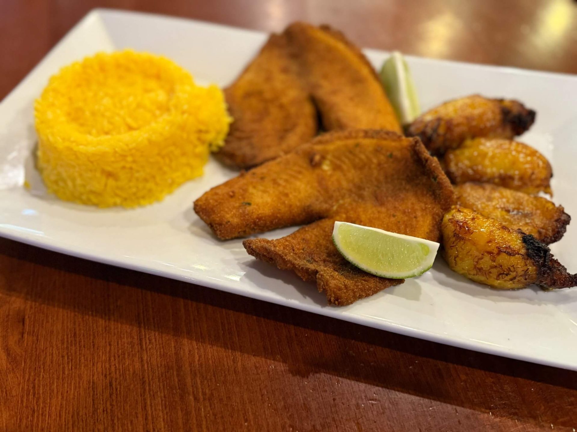 A rectangular plate with yellow rice, fried fish fillets, grilled plantains, and lime wedges on a wooden table.