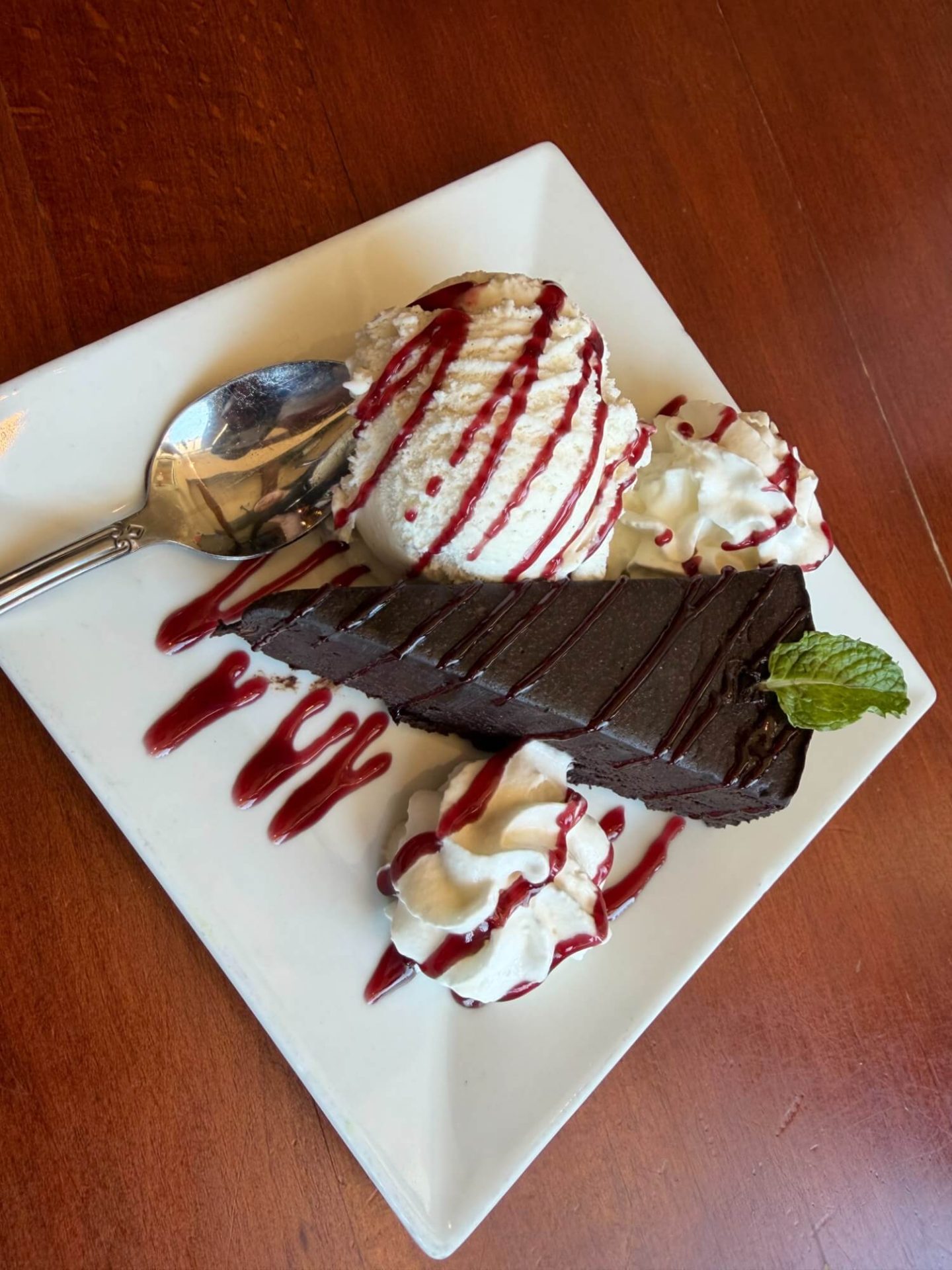 A slice of chocolate cake with a mint leaf, vanilla ice cream, whipped cream, and raspberry sauce on a square white plate with a spoon.