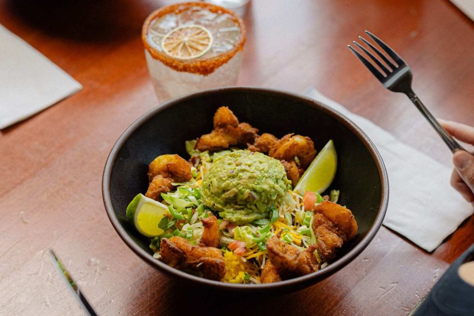 A black bowl filled with shrimp, shredded cheese, lettuce, guacamole, tomato, and lime wedges sits on a wooden table next to a drink and napkin.