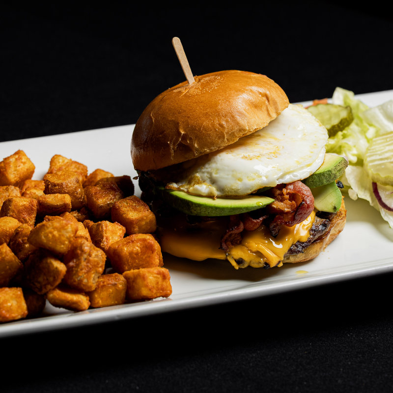Burger with cheese, bacon, avocado, and a fried egg on a bun, served with tater tots, pickles, lettuce, and onion on a rectangular white plate.