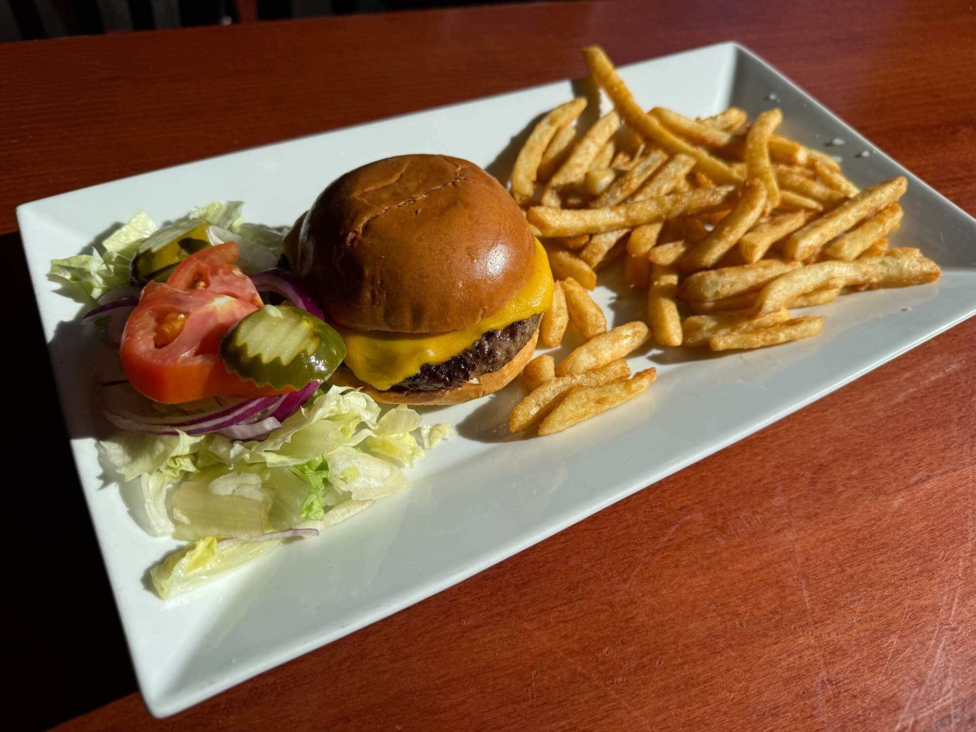 A cheeseburger with lettuce, tomato, onions, pickles, and a side of French fries on a rectangular white plate.