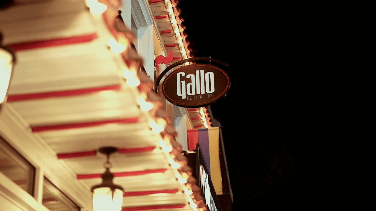 A circular sign with the word "Gallo Restaurant" hangs from the side of a building decorated with string lights at night.