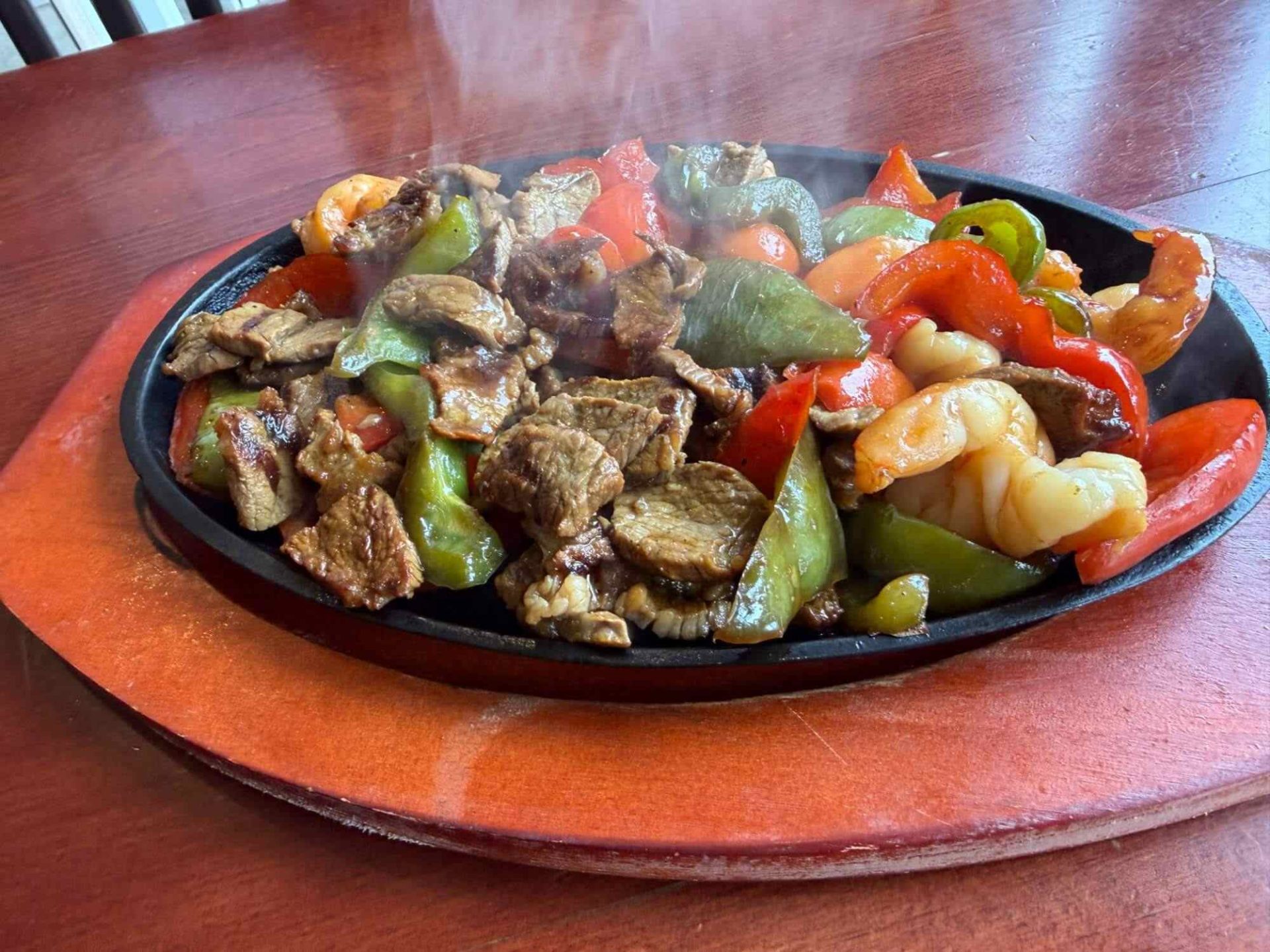 A sizzling platter of beef, shrimp, green peppers, and red bell peppers sits on a wooden table, with steam rising from the food.