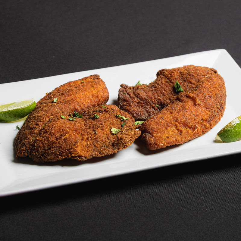 Two breaded and fried fish fillets garnished with chopped herbs, served on a white rectangular plate with lime wedges.