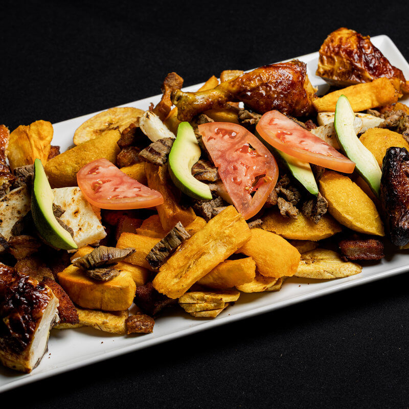 A rectangular plate with fried plantains, yuca, grilled chicken, avocado slices, tomato wedges, and pieces of meat on a black background.