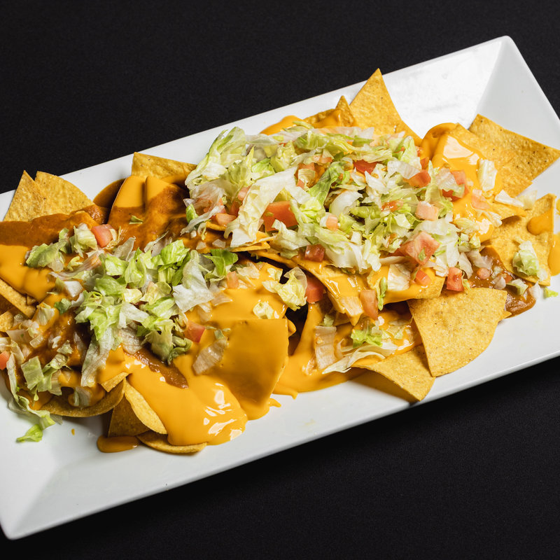 Rectangular white plate with tortilla chips topped with melted cheese, shredded lettuce, diced tomatoes, and other toppings, set on a black background.