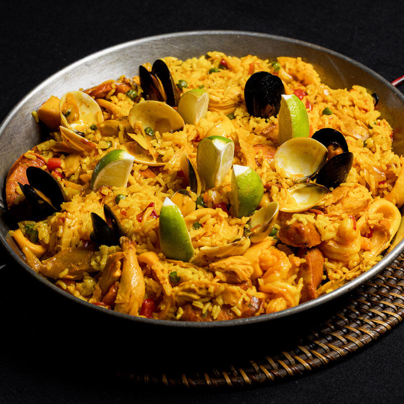 A pan of seafood paella with yellow rice, shrimp, clams, mussels, chicken, and lime wedges, served on a woven mat against a black background.