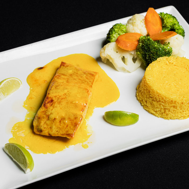 Grilled fish fillet with yellow sauce, served with yellow rice, steamed broccoli, cauliflower, carrot slices, and lime wedges on a rectangular white plate.