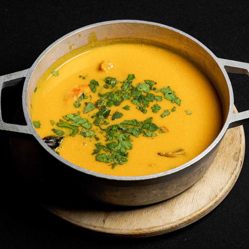 A metal pot filled with orange-colored soup, garnished with chopped green herbs, sits on a round wooden trivet against a black background.