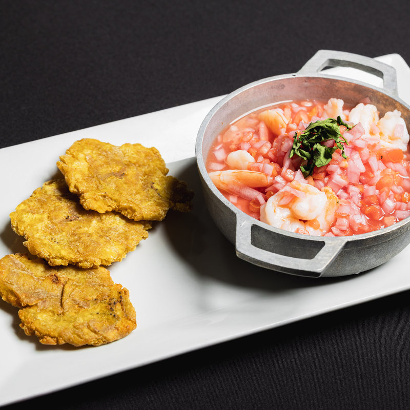 A small pot of shrimp ceviche garnished with herbs is served next to three fried plantain slices on a rectangular white plate.