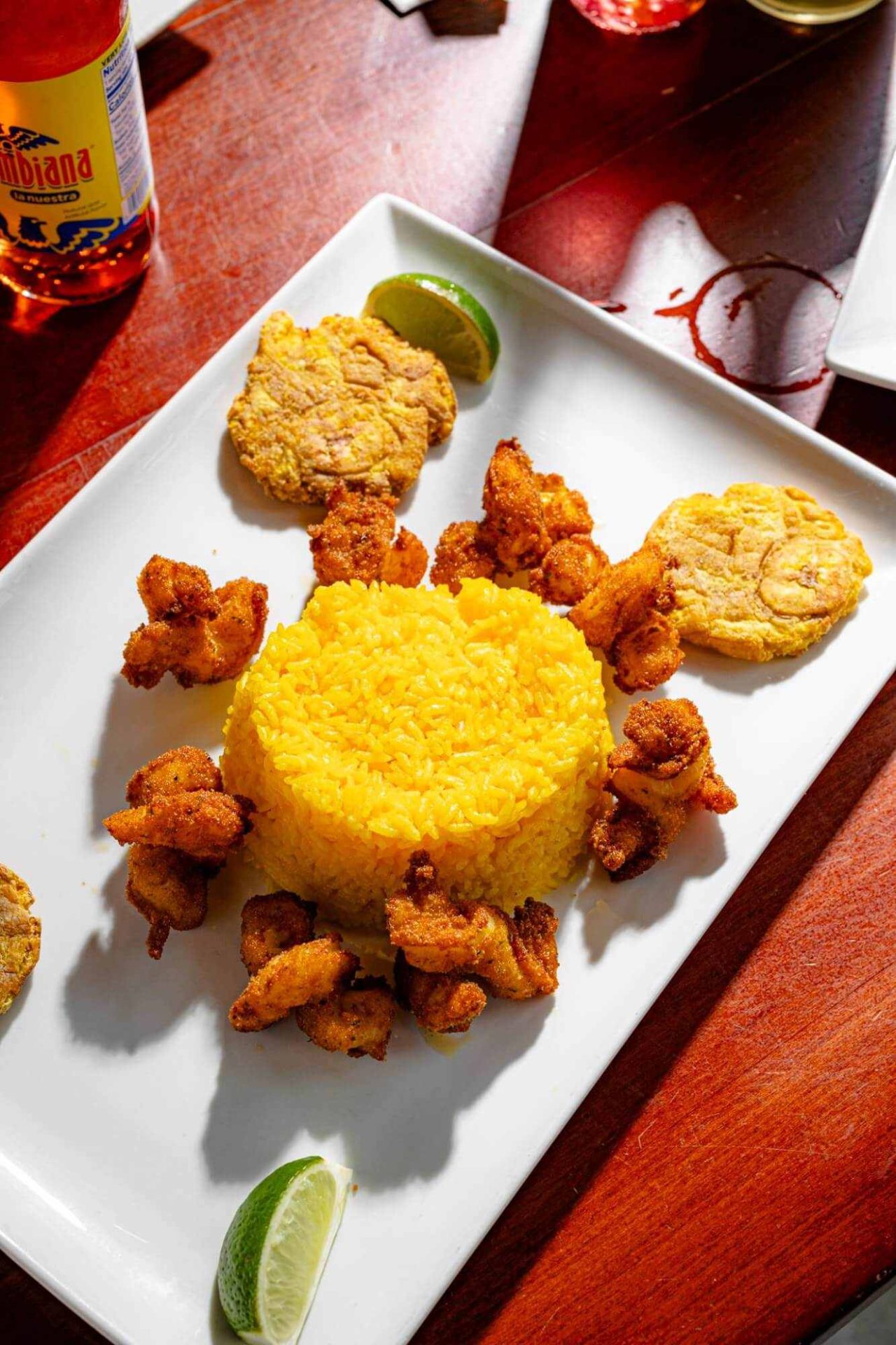 A white plate with yellow rice in the center, surrounded by fried shrimp, two tostones, and lime wedges, on a wooden table.