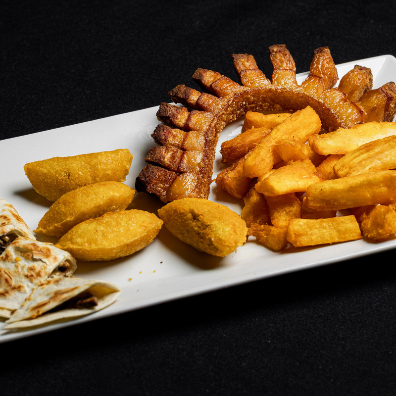 A rectangular white plate with fried pork belly, yellow empanadas, fried cassava, French fries, and pieces of quesadilla on a black background.