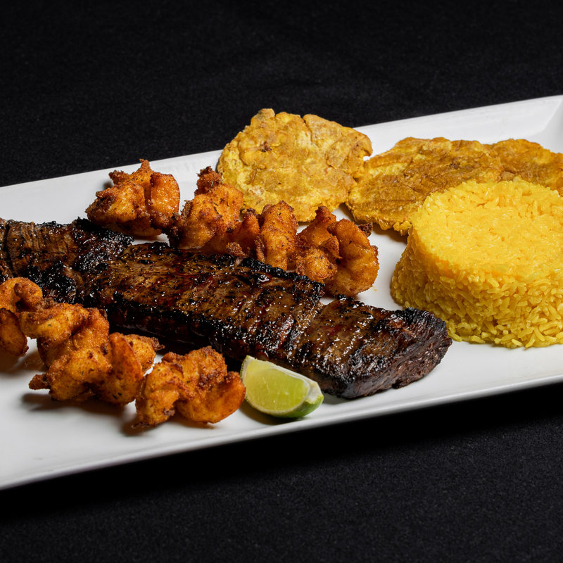 A grilled steak served with yellow rice, fried plantains, fried shrimp, and a lime wedge on a rectangular white plate against a black background.