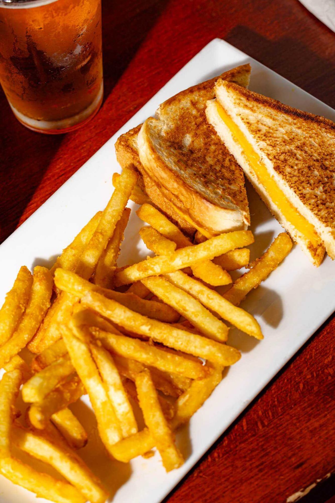A plate with a grilled cheese sandwich cut in half and a serving of French fries, next to a glass of a cold beverage on a wooden table.