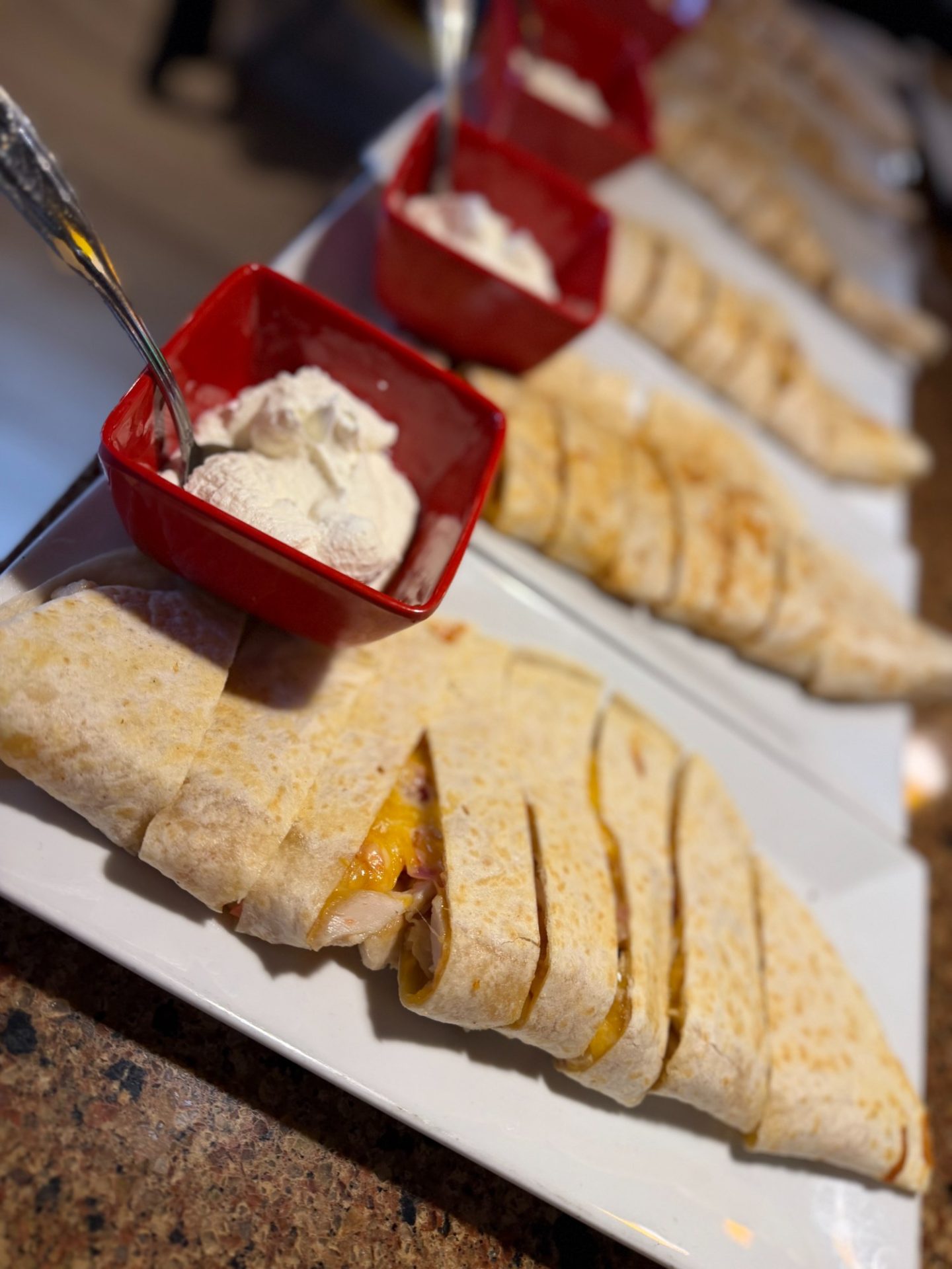 Sliced quesadillas arranged on a white platter, each served with a red bowl of sour cream on a countertop.