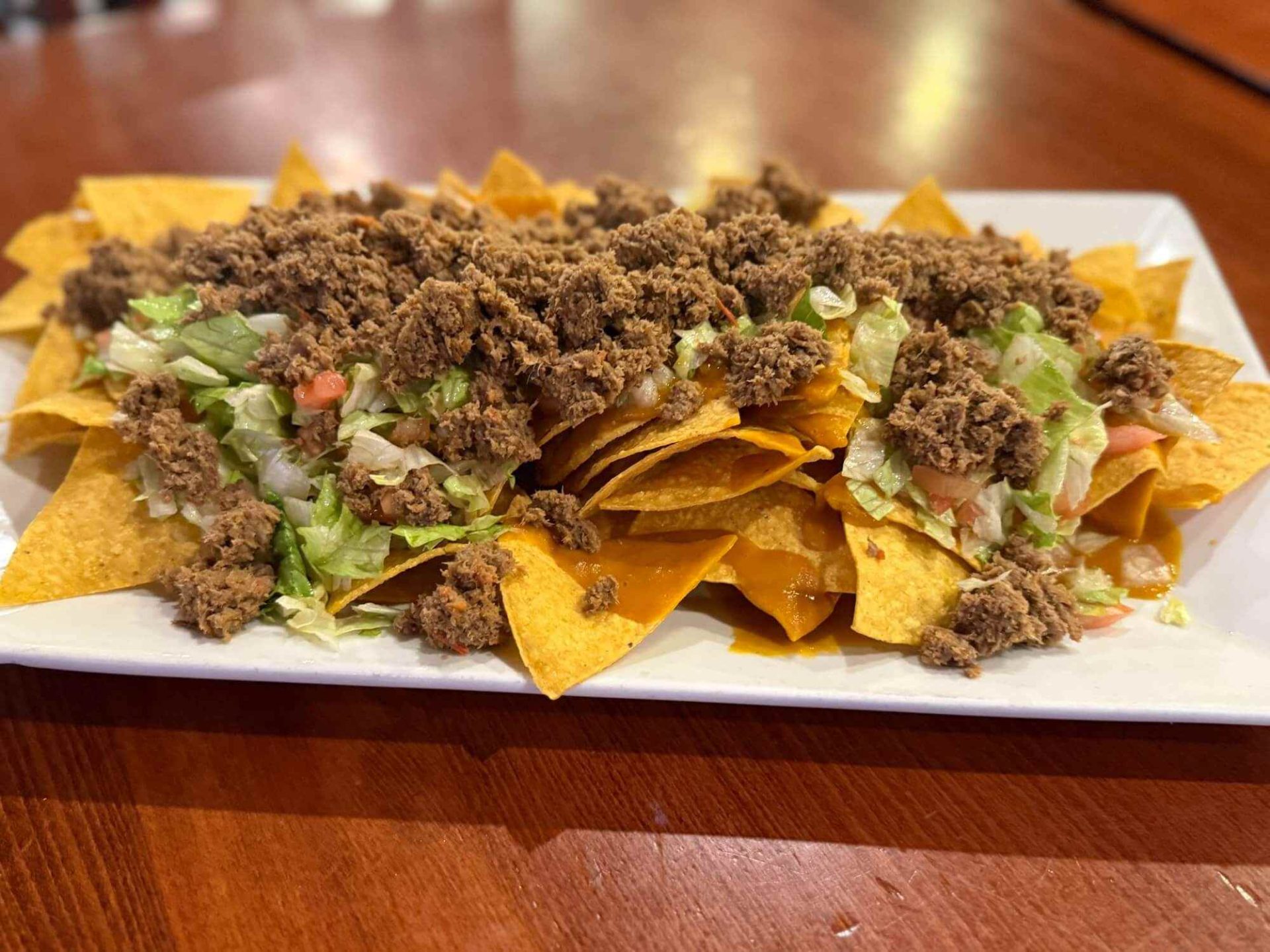 A rectangular plate of nachos topped with ground beef, shredded lettuce, and chopped tomatoes sits on a wooden table.