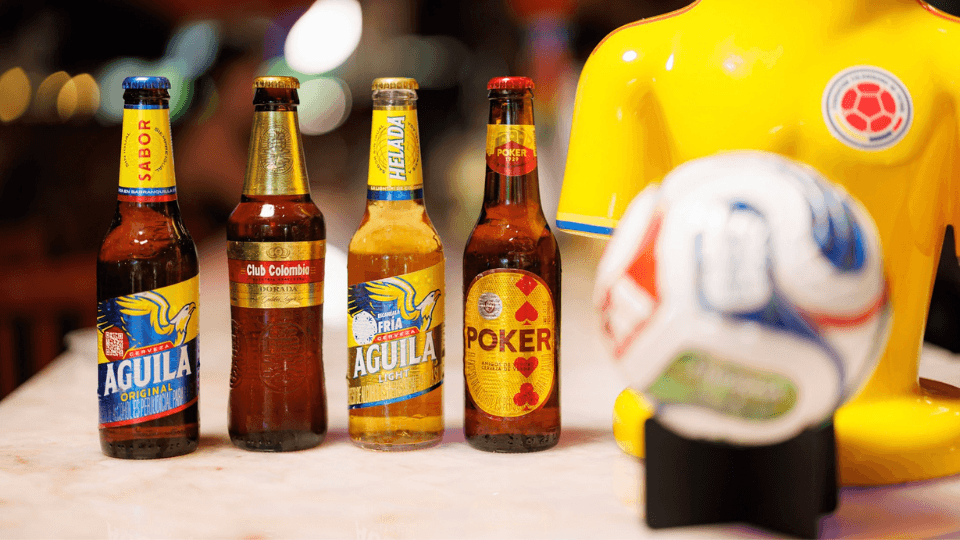 Four Colombian beer bottles—Aguila, Club Colombia, Aguila Light, and Poker—stand on a table beside a yellow soccer jersey and a soccer ball.