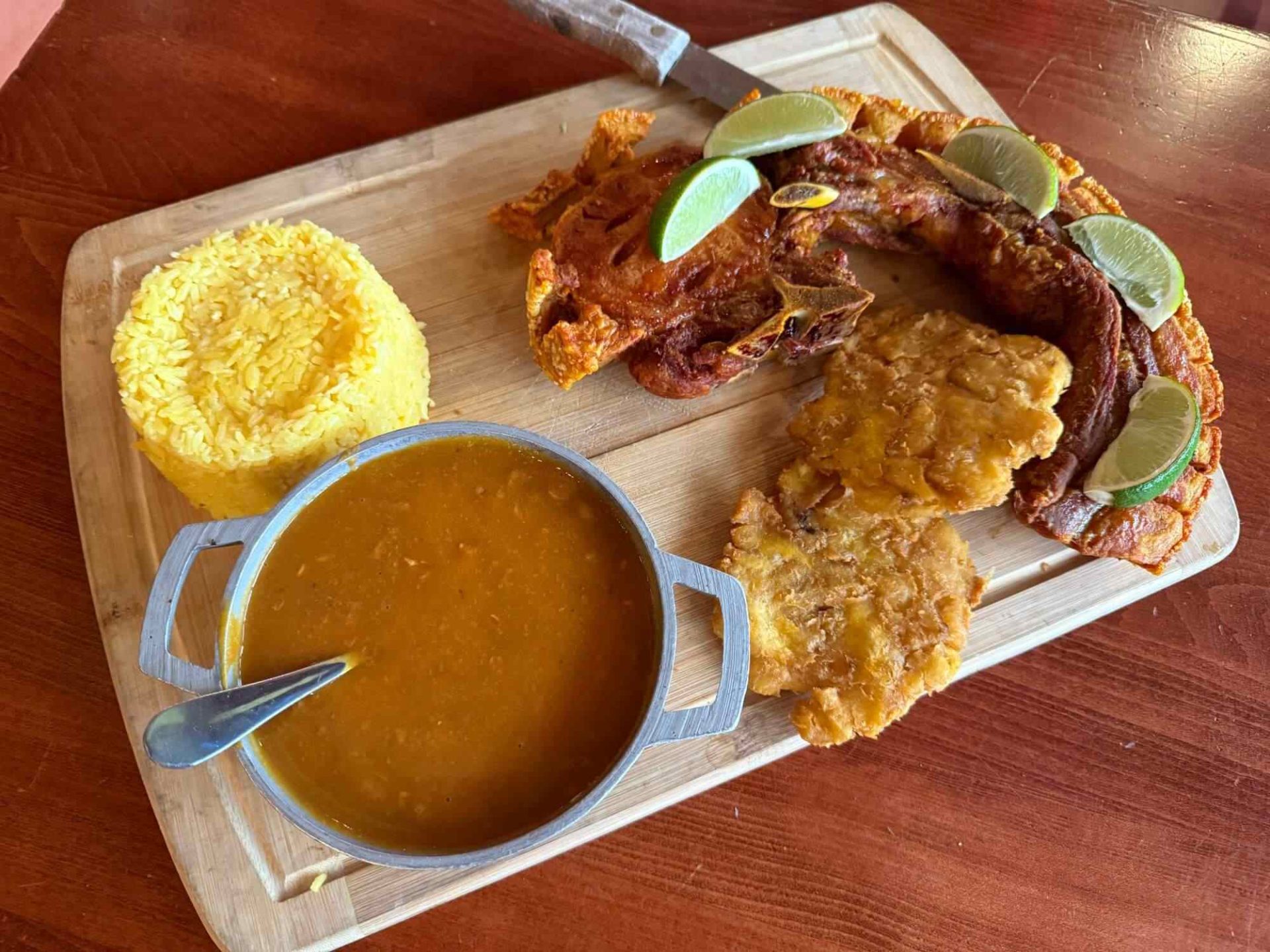 A wooden board with yellow rice, a bowl of bean soup with a spoon, fried pork with lime wedges, and two tostones on a wooden table.