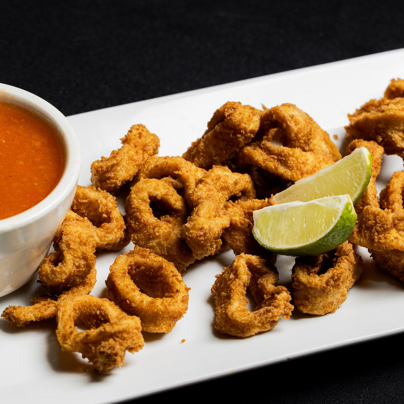 A plate of fried calamari rings with lime wedges, served alongside a small bowl of red dipping sauce on a white rectangular plate.