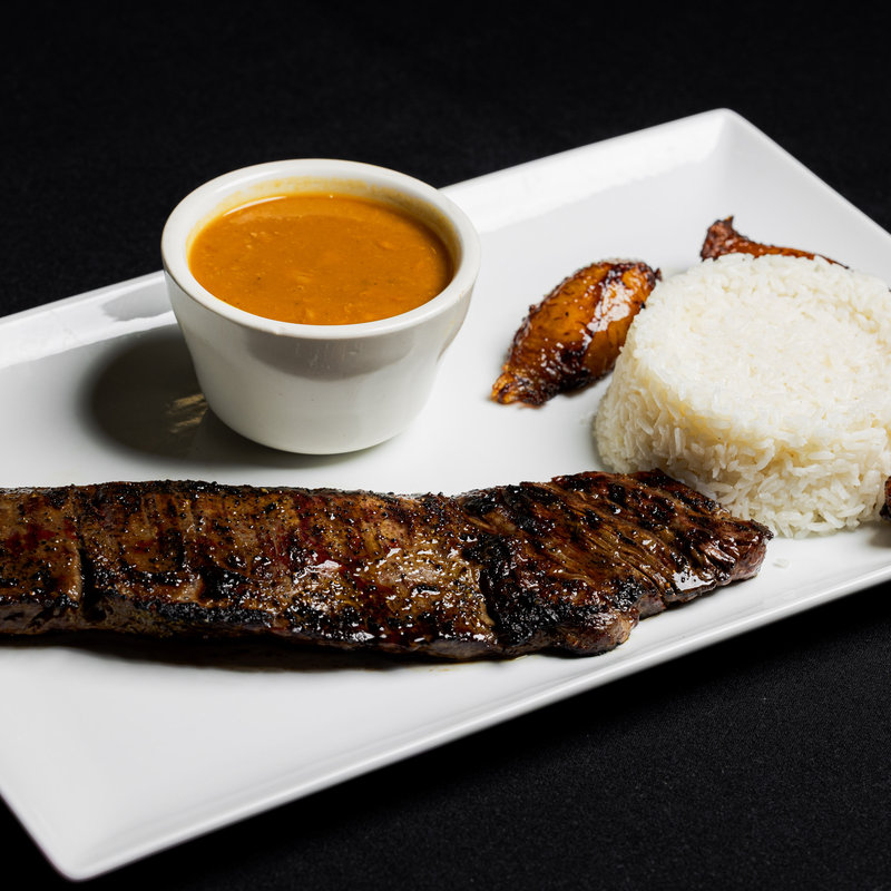Grilled steak, white rice, plantain, and a cup of sauce are served on a rectangular white plate against a black background.
