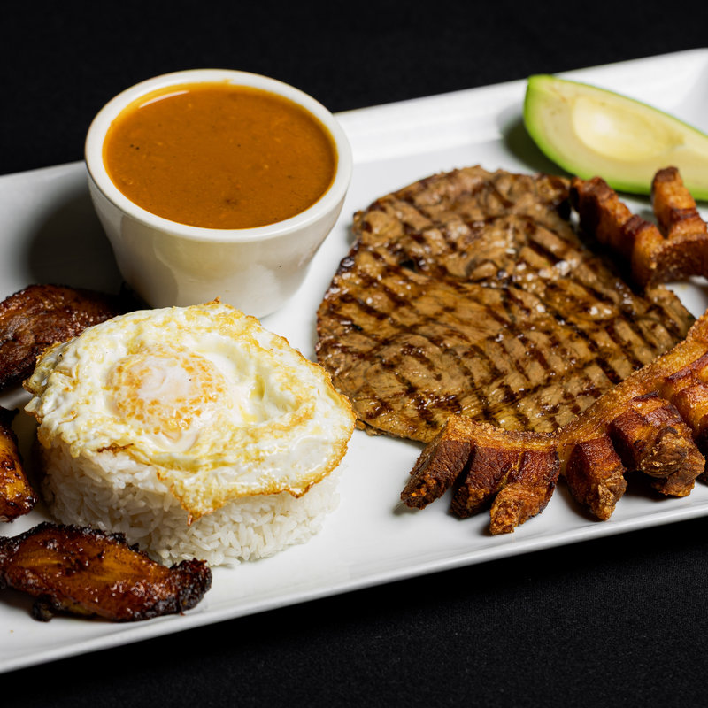 A rectangular plate with grilled steak, white rice topped with a fried egg, fried plantains, avocado slices, chicharrón, and a cup of brown sauce.