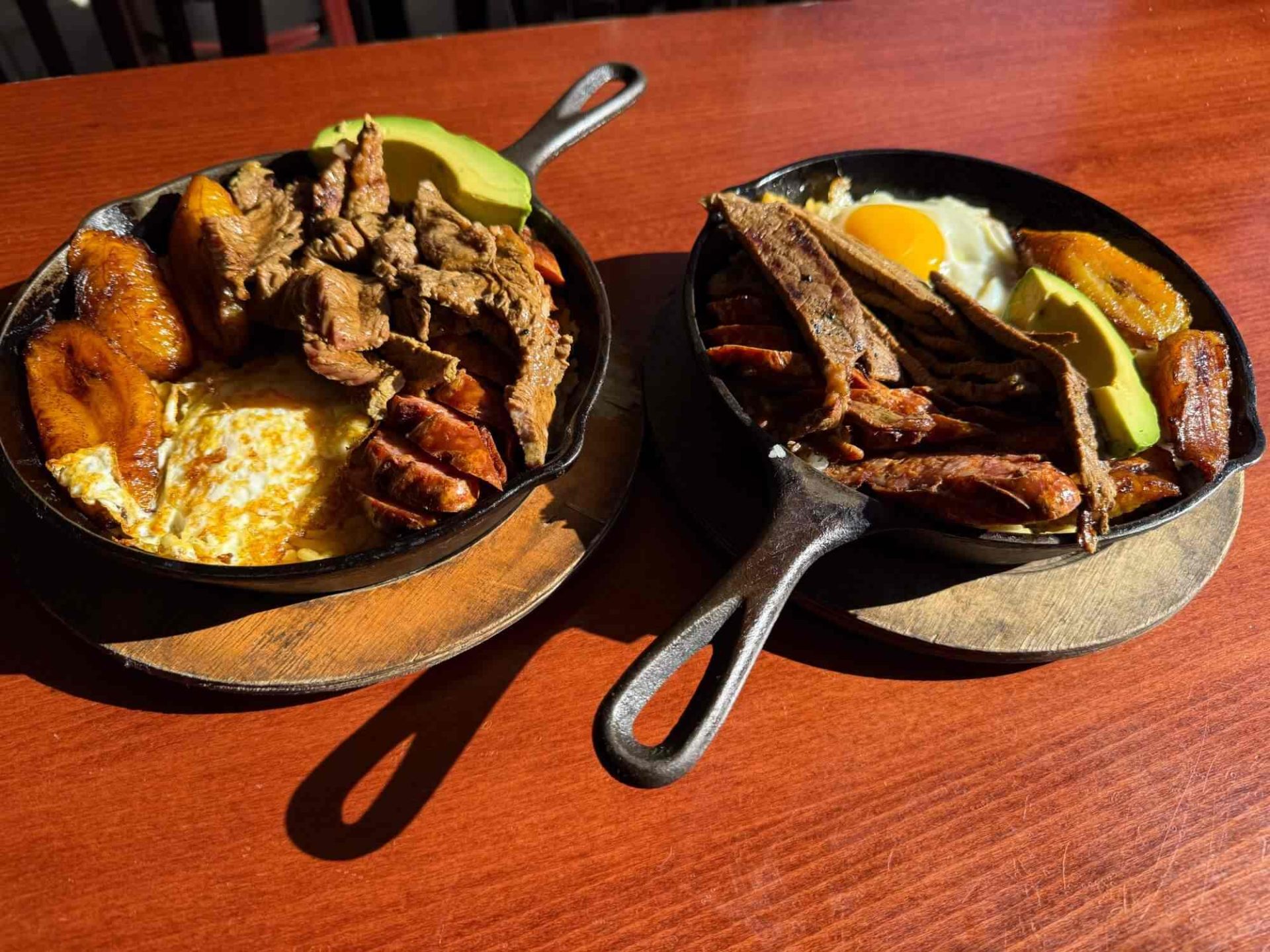 Two cast iron skillets on a wooden table, each filled with grilled meat, fried plantains, avocado slices, and eggs, served on round wooden boards.