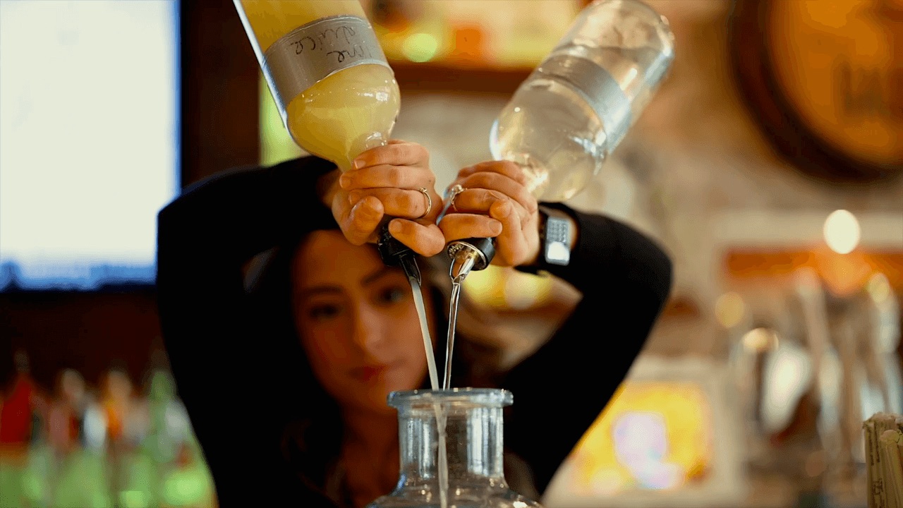 A bartender pours two different liquids from bottles into a large glass container, using both hands to control the flow.