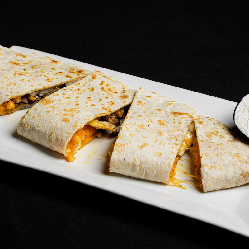 A white rectangular plate holds a sliced quesadilla with visible melted cheese and vegetables, set on a black background.