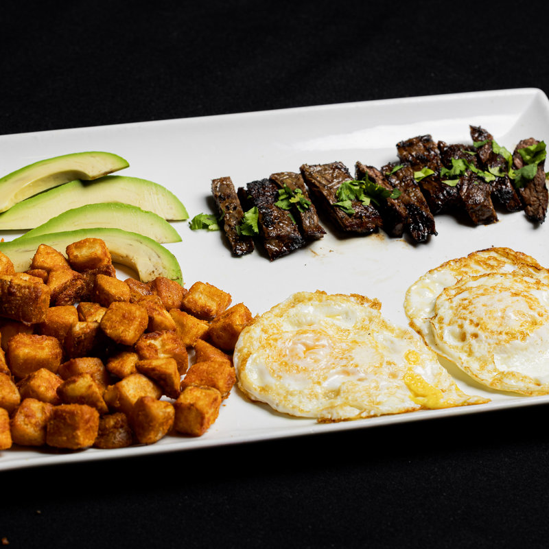 Rectangular plate with sliced avocado, diced potatoes, two sunny-side-up eggs, and grilled steak pieces garnished with herbs, all arranged neatly on a black background.
