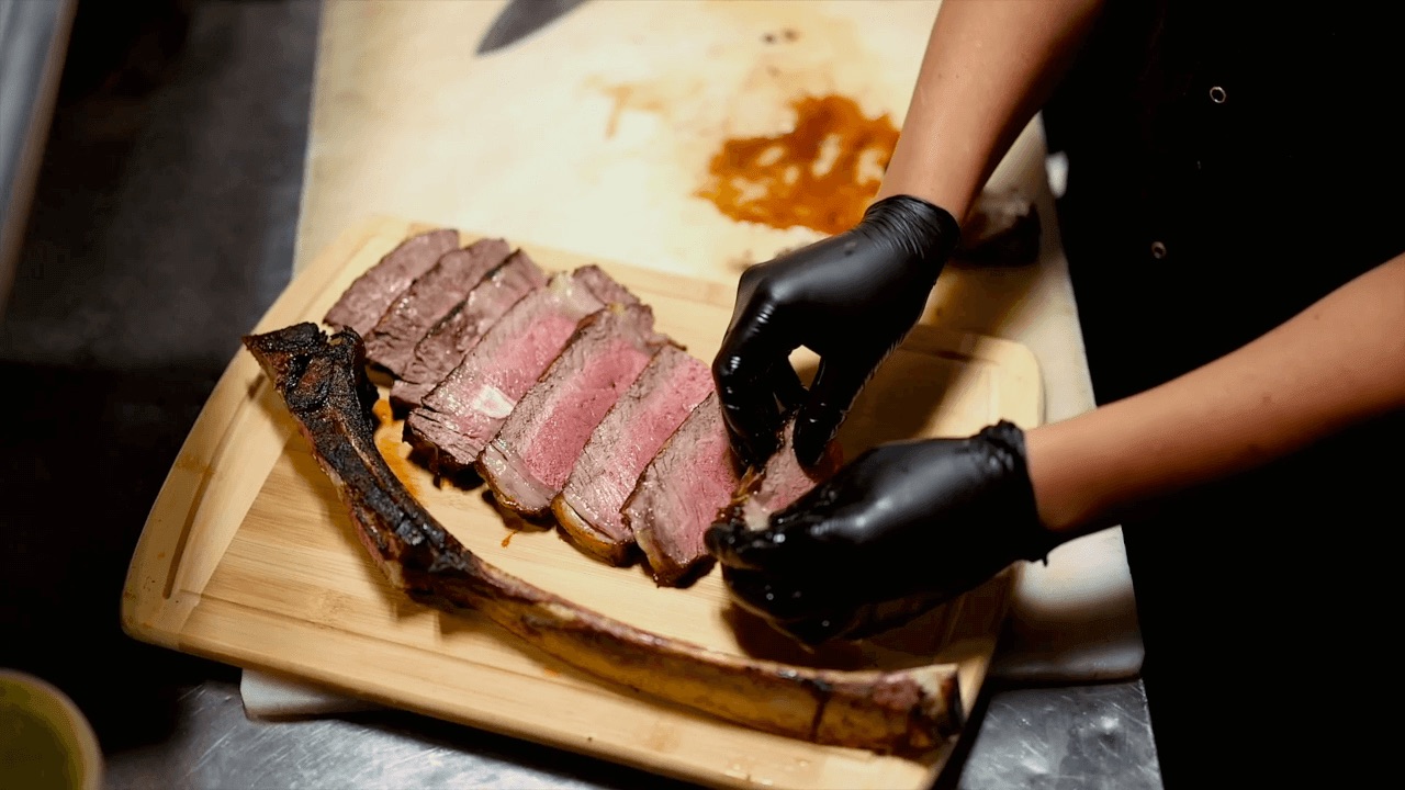 Person wearing black gloves arranges sliced cooked steak on a wooden cutting board, with a large bone on the side.