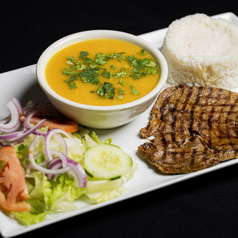 A plate with grilled steak, white rice, yellow soup garnished with herbs, and a side salad with lettuce, tomato, cucumber, and red onion.