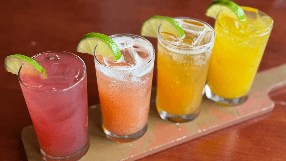 Four colorful cocktails with ice and lime wedges sit in a row on a wooden tray, placed on a reddish-brown surface.