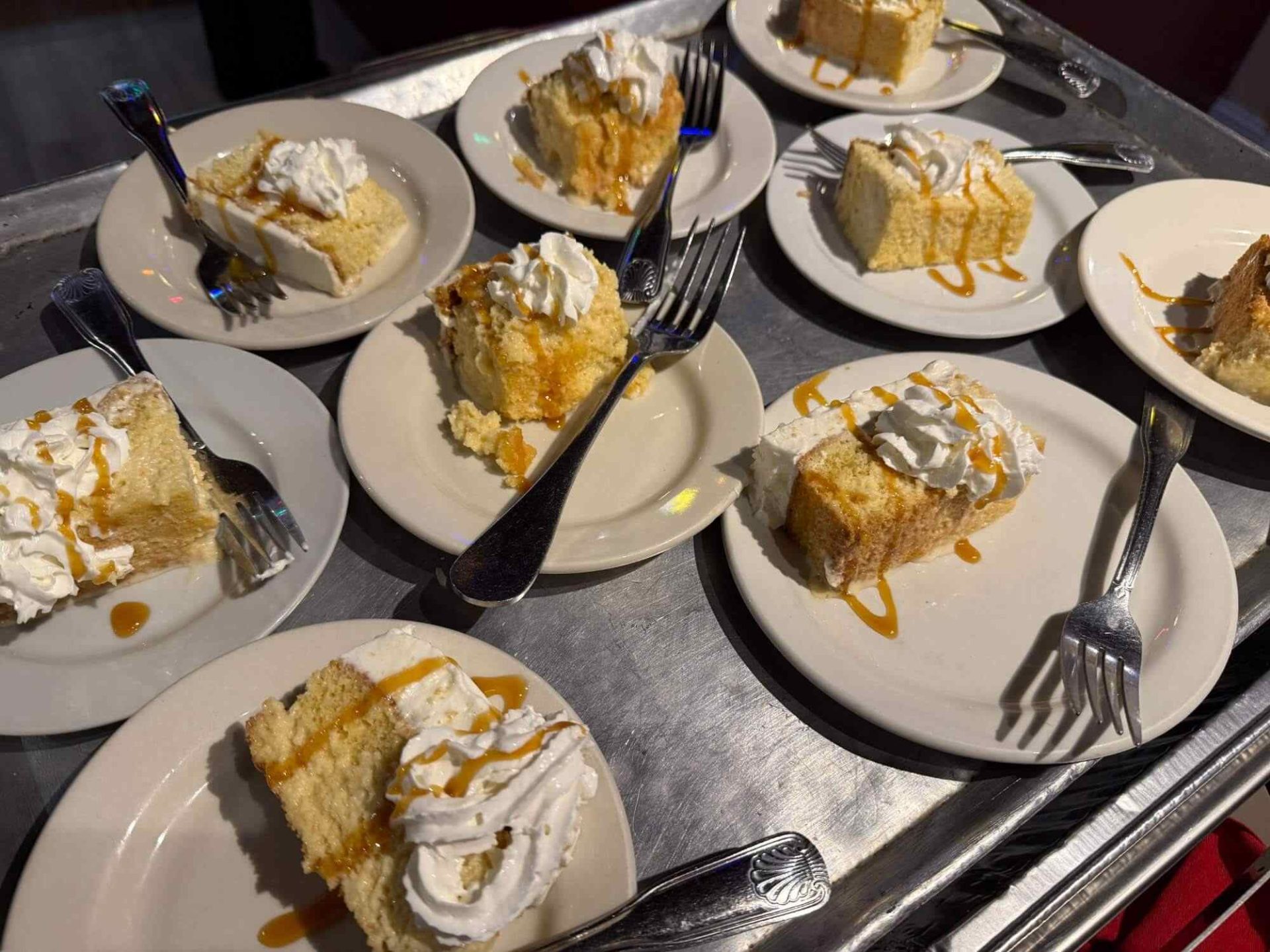 Several plates with slices of cake topped with whipped cream and caramel drizzle, each with a fork, arranged on a metal tray.