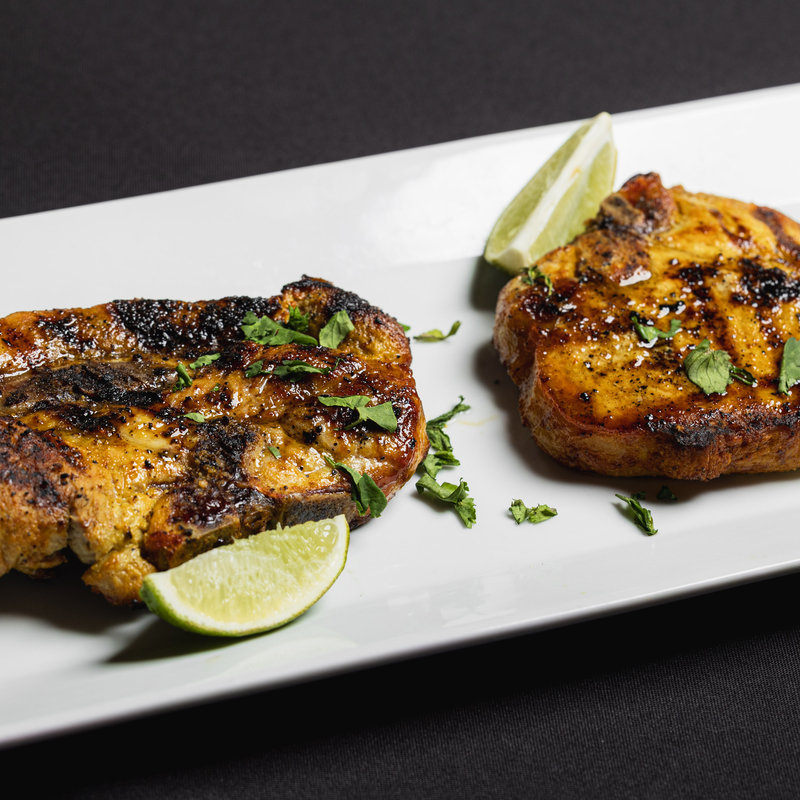Two grilled pork chops garnished with chopped herbs and served with lime wedges on a white rectangular plate against a black background.