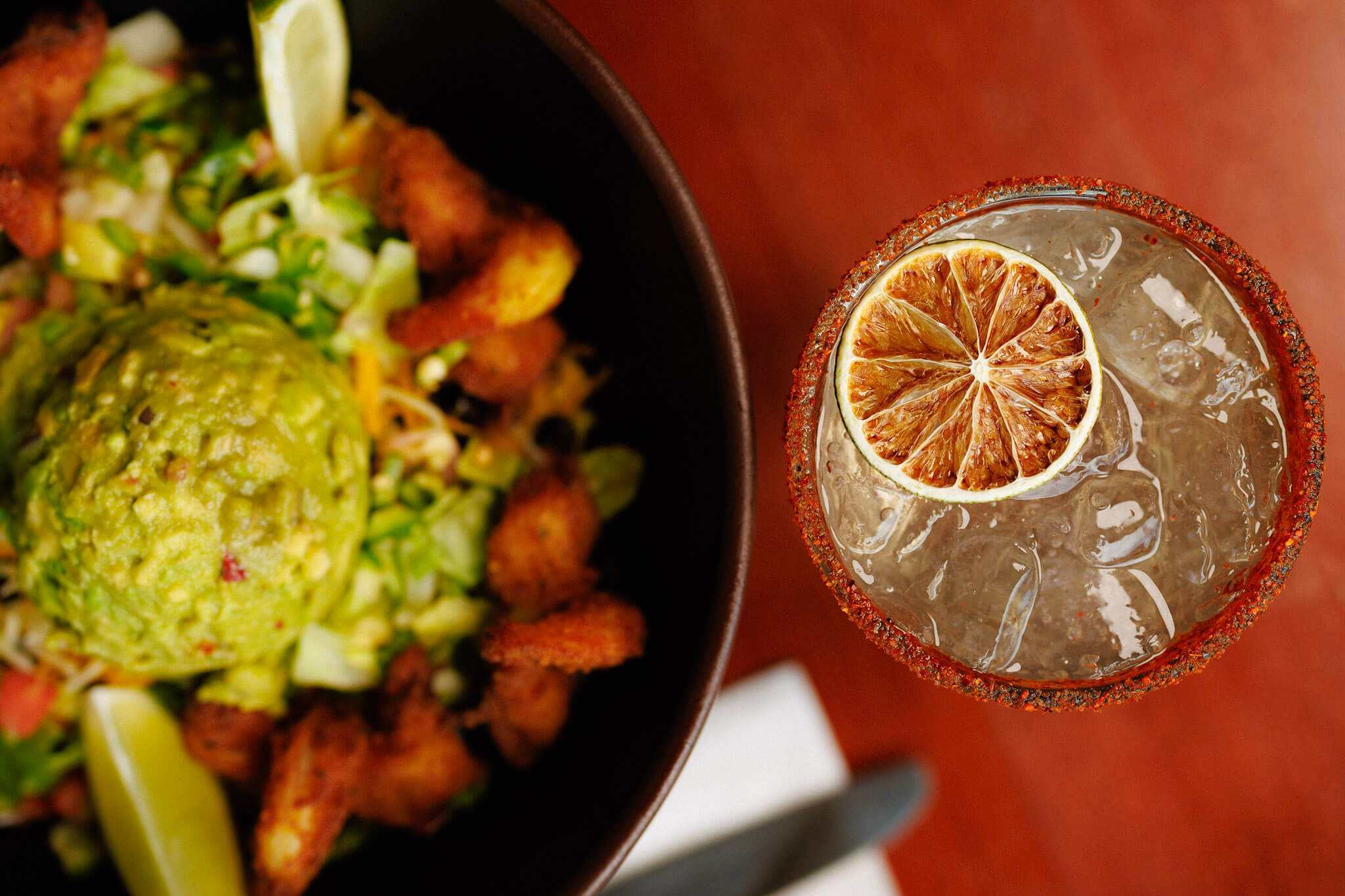 A bowl of food with guacamole and fried shrimp next to a cocktail in a salt-rimmed glass garnished with a dried lime slice.